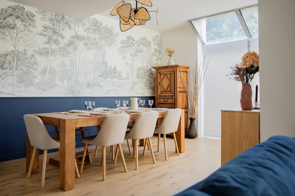 Salle à manger avec grande table en bois, chaises beiges et papier peint forêt, projet Barnabé par Maison Solthée, architecte d'intérieur à Marseille