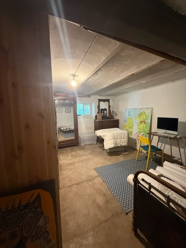 Avant travaux : pièce au sol brut et plafond à poutres, lit en fer forgé et bureau, projet Augustine par Maison Solthée, architecte d'intérieur à Cassis