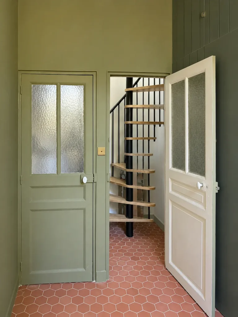 Entrée vert sauge avec porte vitrée, escalier hélicoïdal visible et sol terracotta, projet Augustine par Maison Solthée, architecte d'intérieur à Cassis